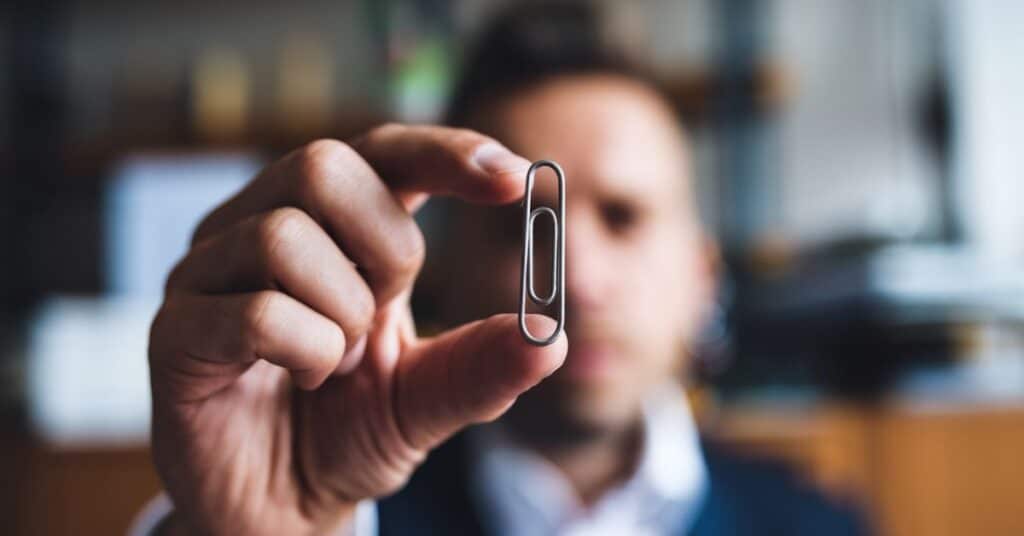 A blurred background of a man's face and his hands are holding a paperclip. The paperclip is straight and silver.