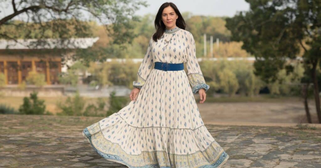 A photo of an adult female standing outdoors. She is wearing a white dress with blue and yellow patterns. The background reveals a landscape with trees and a house.