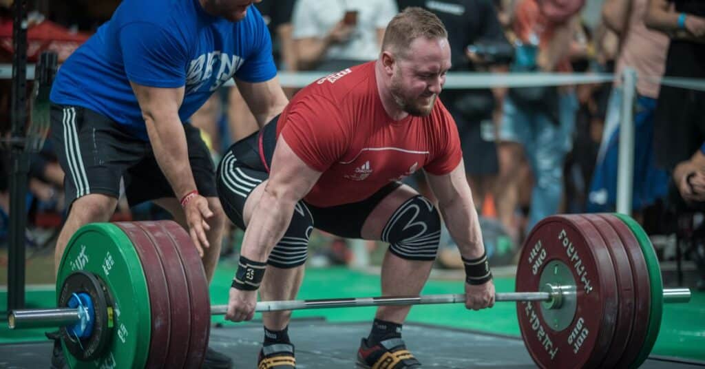 A man is trying to lift a heavy barbell with the assistance of a spotter. The man is wearing a red shirt and the spotter is wearing a blue shirt.