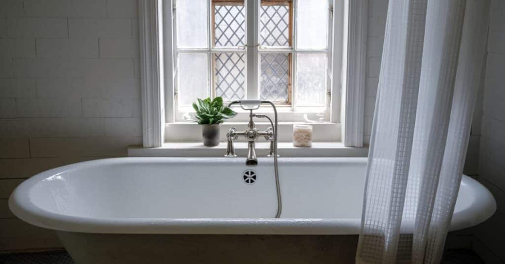 A photo of a bathtub in a bathroom. There is a white curtain near the bathtub and it has a window with a small plant.