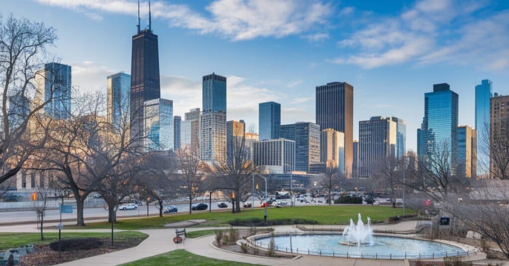 A panoramic view of Chicago's skyline. The foreground reveals a city park with a walking path, trees, and a fountain. The background contains a multitude of skyscrapers.