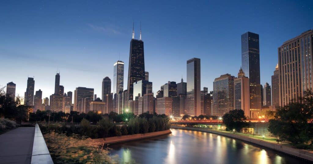 A photo of the Chicago skyline at night, with the city's skyscrapers illuminated by bright lights. The river snakes through the city, reflecting the lights and buildings. The foreground contains a pathway and plants.