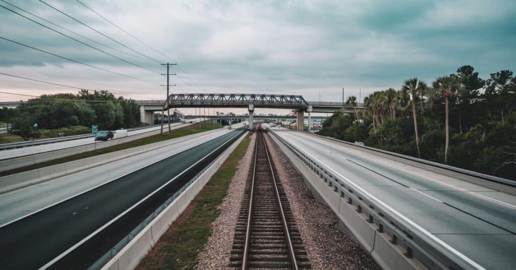 A photo of Florida's transportation infrastructure. There is a highway with multiple lanes, a railway track alongside the highway, and a bridge in the background.