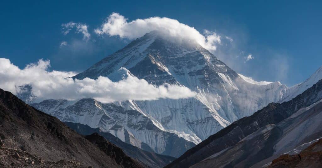 A photo of Everest from the base, with the peak shrouded in clouds. The mountain is surrounded by clear blue skies, with a few mountains in the background.