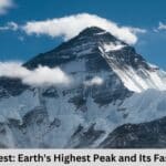 A photo of Mount Everest with a clear blue sky and a few clouds. The peak is capped with snow and is partially shrouded in clouds.