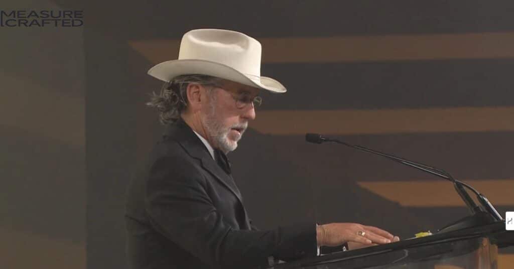 An image of Arch Aplin while giving Acceptance Speech in 2023. He is weairng a black suit with white hat.