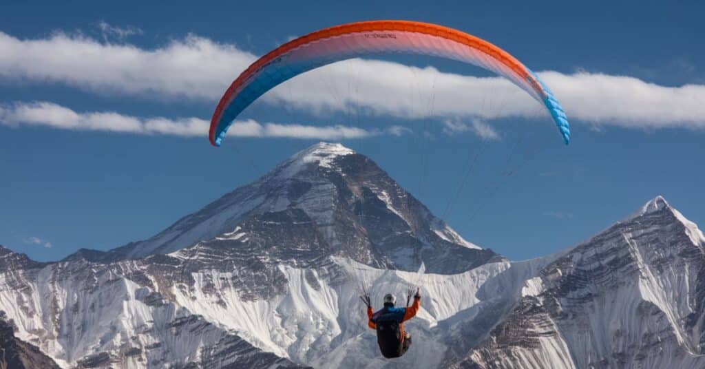 A photo of a paraglider flying over the mountains. The paraglider is wearing a bright orange and blue suit and a helmet. The background reveals the vast expanse of the mountain range. The sky is clear with a few clouds.