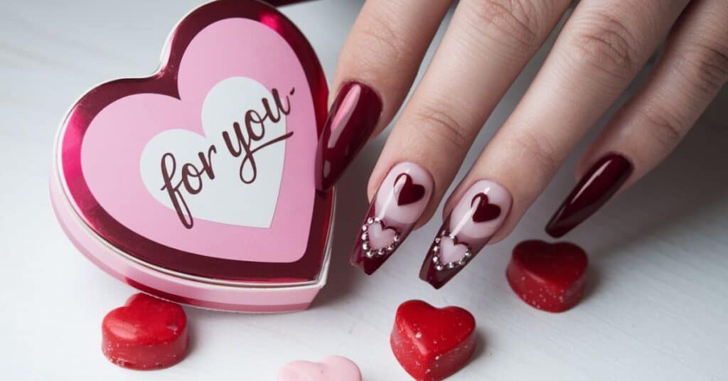 A photo of a set of coffin shaped nails with Valentine's Day theme. The nails have a heart shaped design and are dipped in a dark red polish. There is a pink and red heart shaped box with the words "For You" written on it. There are also few heart shaped candies.