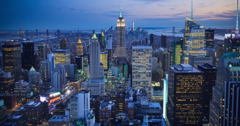 A photo of an aerial view of NYC at night. The city is illuminated with bright lights, showing the Empire State Building, Times Square, and other iconic buildings. The Hudson River reflects the city lights.