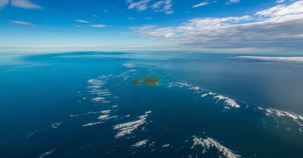 An image of the middle of the Central North Sea during the day. The sea is a deep blue, with scattered white foam from the waves. In the middle of the sea, there is a small, green area.