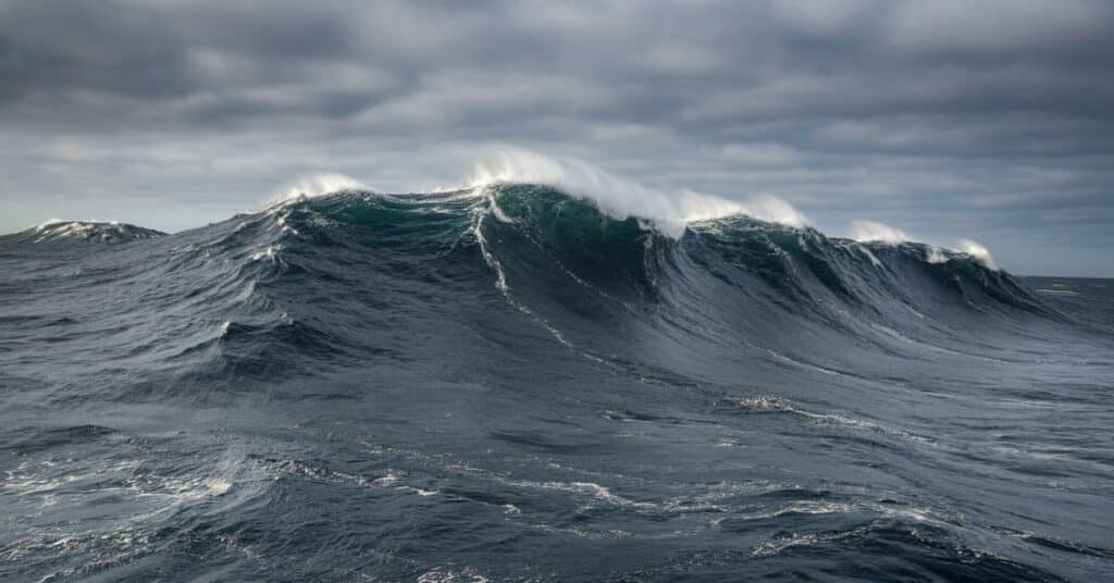 A photo of the middle of the ocean with massive waves. The waves are towering over the ship and are white at the top. The sky is overcast, with dark clouds.