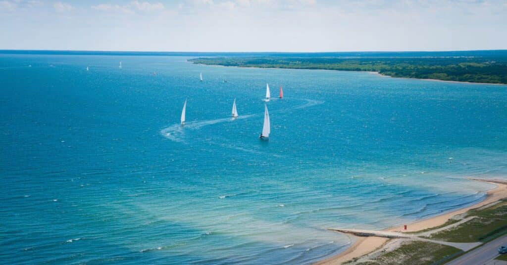 A photo of the sparkling blue waters of Lake Michigan with a few sailboats gliding across it. The shoreline has sandy beaches and a paved road.