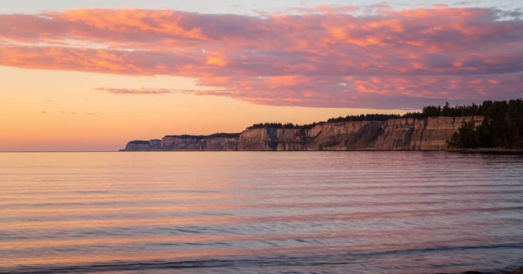 A photo of Lake Michigan at sunset. The lake is calm, with the water reflecting the vibrant orange and pink sky. In the background, there are cliffs with a few trees.