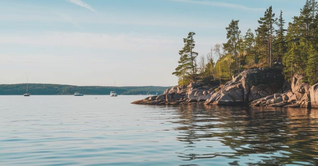 A photo of Lake Michigan with a clear sky. The water is calm, reflecting the sky and the surrounding landscape. The shoreline is rocky, with trees growing out of the rocks. There are a few boats anchored in the lake.