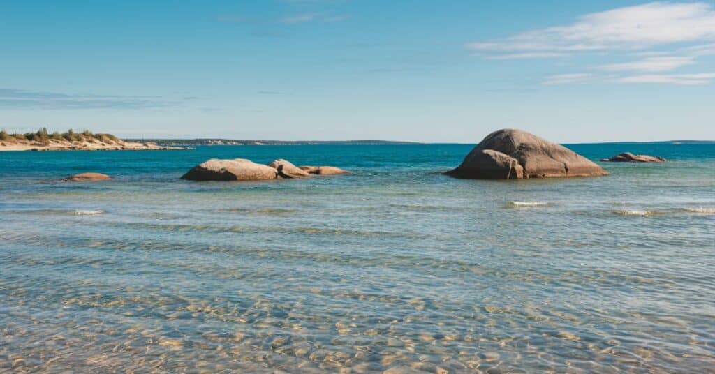 A photo of the clear blue water of Lake Michigan with a few large rocks in the middle of the lake. The water is calm, with only a few small waves. The shoreline is lined with sand dunes.