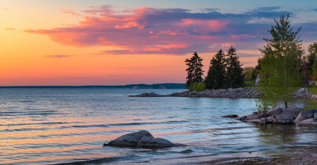 A photo of the lake at sunset. The sky is painted with shades of orange, pink, and blue. The water is calm, reflecting the sky. There are rocks and a tree near the water. The shoreline is rocky and has a few trees.