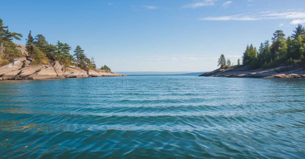 A serene, medium shot of lake with clear, blue water. The surface of the water is calm, with only a few small waves. The sky above is clear, with a few clouds. The shoreline is rocky and has a few trees.