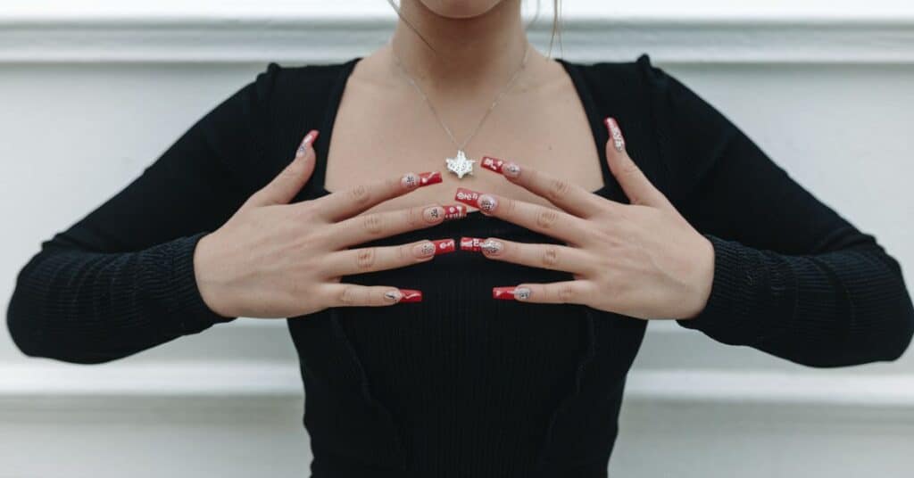 A medium shot of a woman with long, red nails standing in front of a white background. She is wearing a black dress and a necklace.