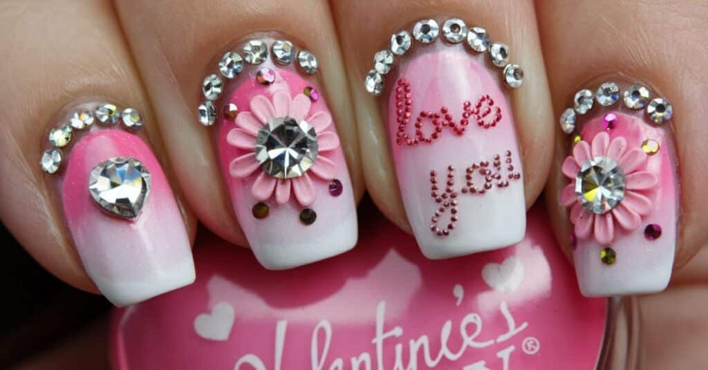 A close up shot of a hand with beautiful rhinestones and accents designed for valentine's day nails. There are hearts, flowers, and the words "Love You" written in cursive. The nails have a pink and white gradient.